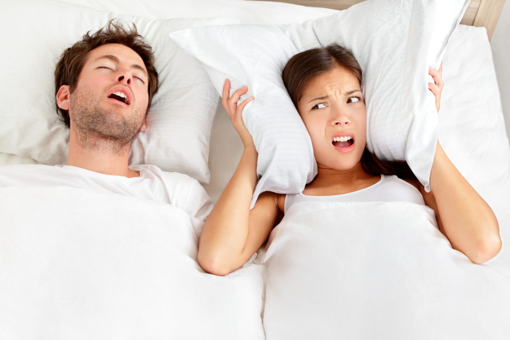 . Couple in bed, man snoring and woman can not sleep, covering ears with pillow for snore noise. Young interracial couple, Asian woman, Caucasian man sleeping in bed at home.